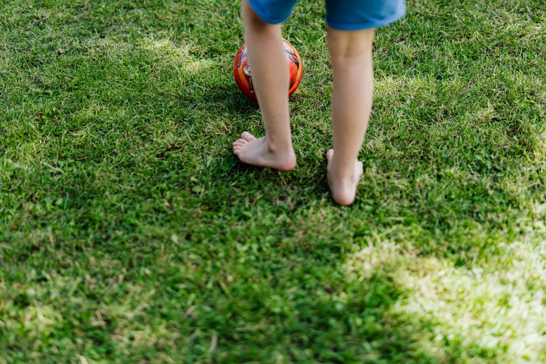 A child playing barefoot with a soccer ball on a grassy field in daylight.